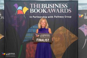 Image shows: Lucinda standing holding a sign with the text 'The Business Book Awards 2025 Finalist'